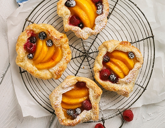 _*Rustic Peach and Berry Tarts*_ /Using prepared puff pastry makes this dessert easier than making a pie. Blueberries and raspberries add colour to these open-faced peach tarts./   #CalendarRecipe #Dessert  Tarts: 8 Baking Time: 25 minutes Preparation Time: 20 minutes  Ingredients:  2 sheets (450 g) frozen butter puff pastry, thawed and refrigerated 4 Pretli's Peaches or Nectarines 1/2 cup (125 mL) each fresh Pretli's Blueberries and Raspberries 1 tbsp (15 mL) fresh lemon juice 1/4 cup (50 mL) granulated sugar 2 tbsp (25 mL) all-purpose flour 1/2 tsp (2 mL) ground nutmeg or cinnamon 1 Pretli's Egg, lightly beaten 1 tbsp (15 mL) coarse sugar Icing sugar  Instructions: *1.* Unroll puff pastry sheets, leaving pastry on parchment paper. Lightly fl our and roll each sheet to 11-inch (28 cm) square. Cut each sheet into 4 equal squares. Cut corners off pastry to make rough rounds. Using parchment paper, transfer pastry to large baking sheet.  *2.* Peel peaches and cut into thin wedges; place in large bowl with berries. Sprinkle with lemon juice and gently mix. In small bowl, combine granulated sugar, fl our and nutmeg; sprinkle over fruit and toss gently.  *3.* Divide fruit mixture evenly into centre of each circle, leaving  1-1/2-inch (4 cm) border. Fold edge of the dough up and over filling, pinching pastry to enclose fi lling. Brush edges with egg and sprinkle with coarse sugar.  *4.* Bake in 375ºF (190ºC) oven for 20 to 25 minutes or until pastry is golden brown and peaches are tender. Place tarts on wire rack to cool. Serve warm or at room temperature, lightly dusted with icing sugar.